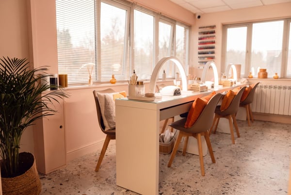 The nail studio at NU Beauty in Haverhill, a long white communal nail table with arched LED lamps running toward the windows, ready for gel manicures