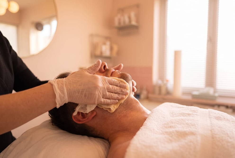 A close, warm detail shot in the NU Beauty facial room in Haverhill: a gloved therapist hand smooths a warm cleansing cloth across a client's jaw and neck, an illustration of the cleansing step of a facial