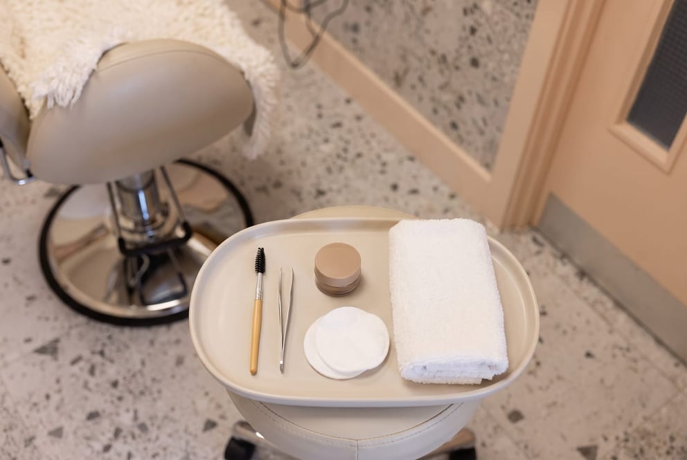 A tidy top-down still-life on a pale tray in the brow area at NU Beauty: a clean spoolie brush, a small pot of brow tint, folded cotton pads and a soft towel, laid out ready for a gentle brow tidy and tint