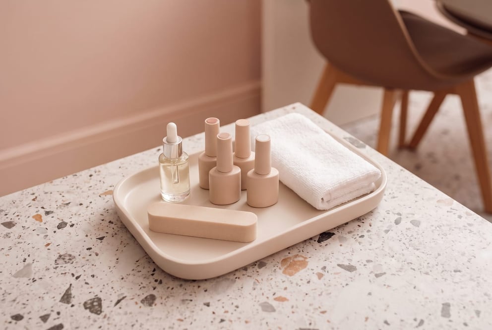 A calm flat-lay on a pale tray in the nail studio at NU Beauty: a small glass bottle of cuticle oil with a dropper, a few plain unbranded gel polish bottles with their labels turned away, a soft buffer and a folded white towel, arranged with plenty of space around them