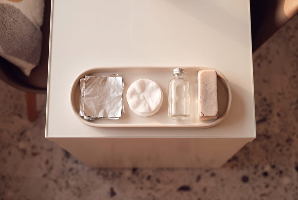 A neat top-down still-life on a tray in the nail studio at NU Beauty: squares of foil, folded cotton pads, a plain unlabelled bottle turned away from the camera and a soft nail buffer, laid out ready for a gentle acetone soak-off removal