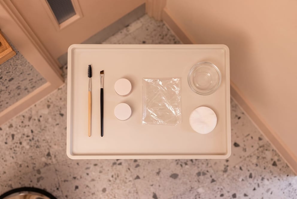 A still life on a cream tray in the brow area at NU Beauty: a slim brow spoolie and an angled brush beside two small unbranded pots of lamination cream, a folded square of clear film, a little glass tint bowl and a soft cotton pad, with the salon's warm styling shelf behind