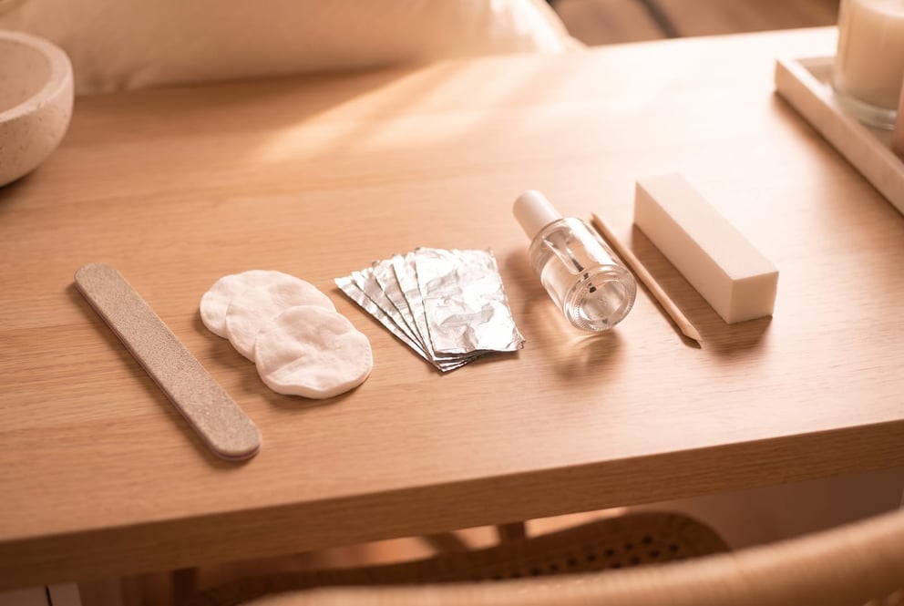 A calm top-down tray of the kit for soaking acrylic nails off: a coarse file, folded cotton pads, squares of foil, a small unlabelled bottle and a wooden cuticle pusher