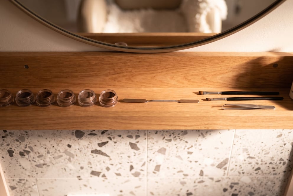 A top-down flat lay of HD brow tools on a dark surface at NU Beauty in Haverhill: small pots of custom-blended brow tint, a mixing spatula, angled brushes, tweezers and a brow mapping pencil